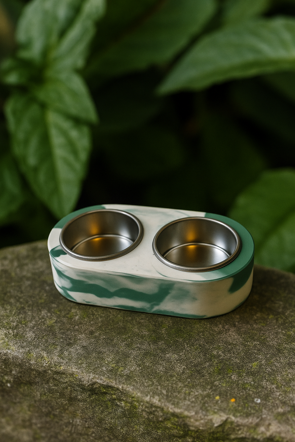 Two-compartment metal feeder with green and white pattern on a stone surface with leaves in the background